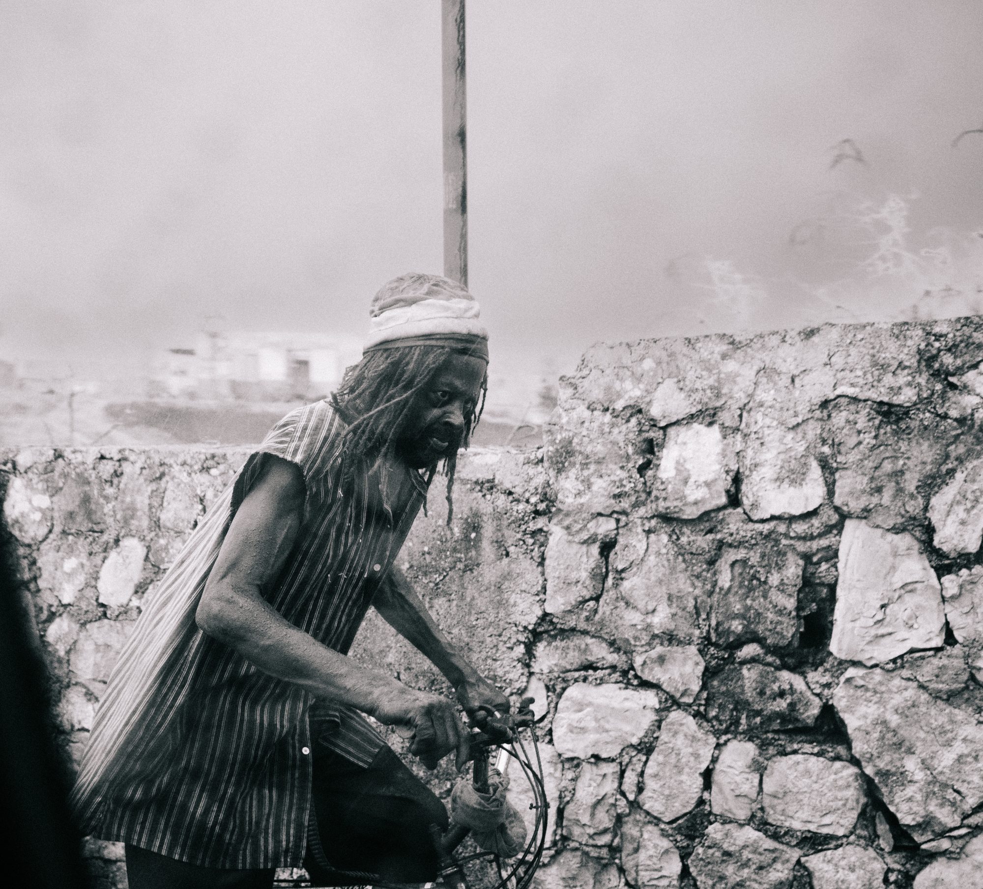 Man with a head wrap on a bicycle by a stone wall, black and white