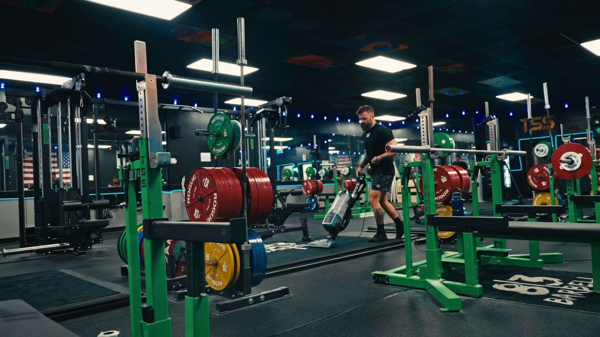 A bearded man in a black T-shirt vacuums the floor at 813 Barbell between green squat racks loaded with red, yellow and blue plates