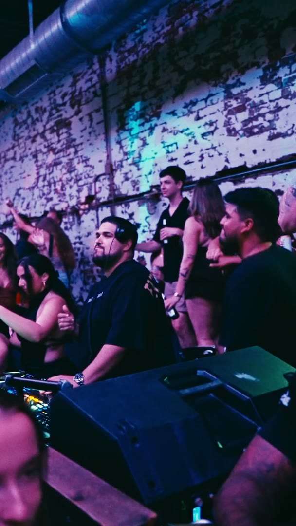 A DJ in headphones at the decks with a crowd dancing behind him against a painted brick wall