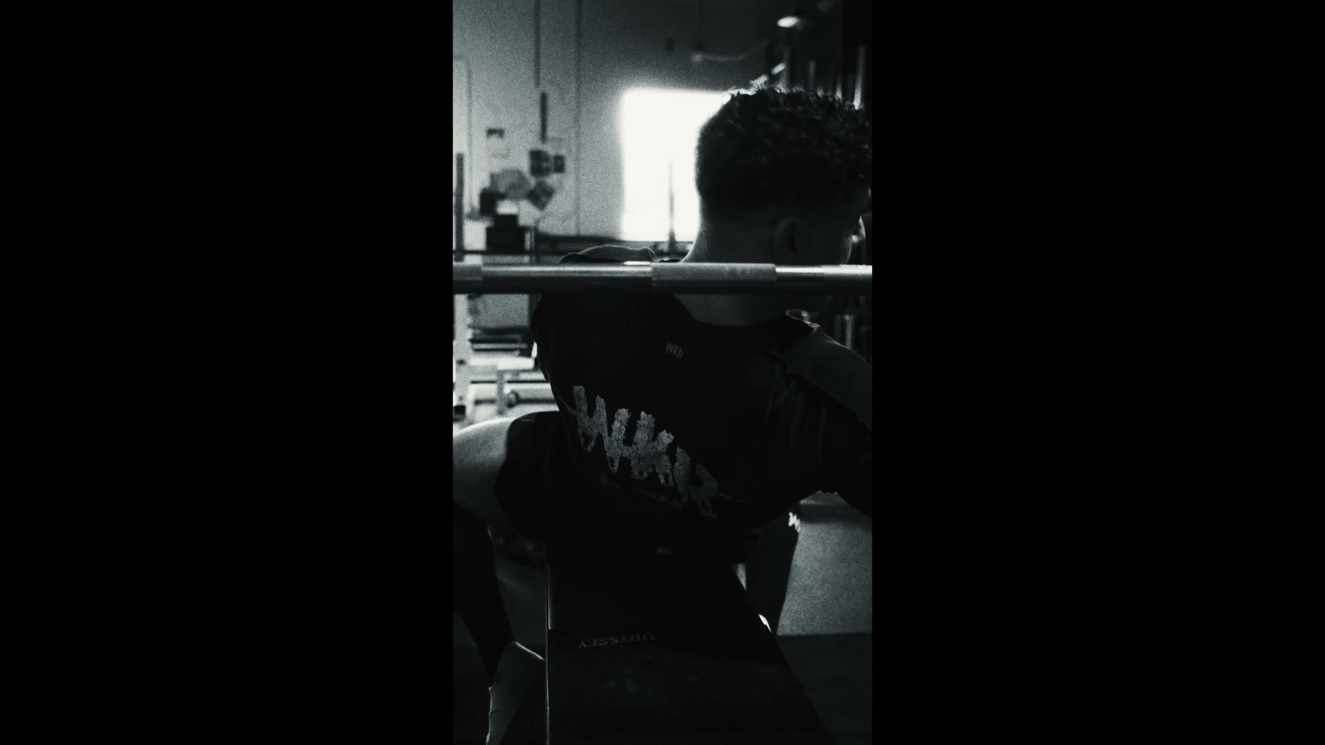 Black and white shot from behind of a man with a barbell across his shoulders in a gym, a bright window in the background