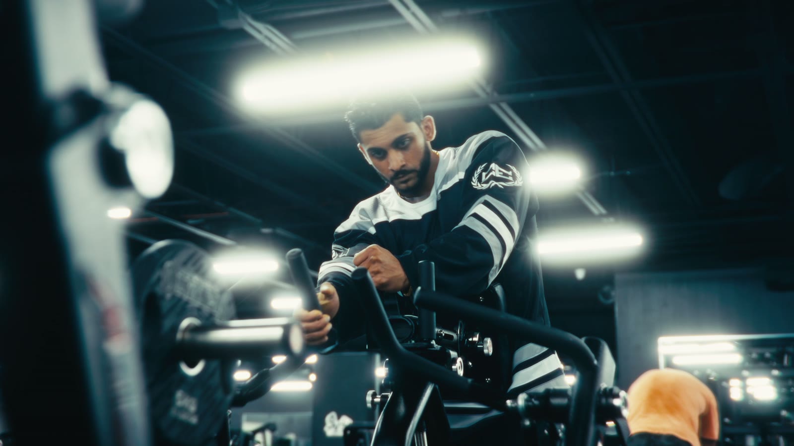 A bearded man in a black and white striped jersey loads a plate machine in a dark gym under bright overhead lights