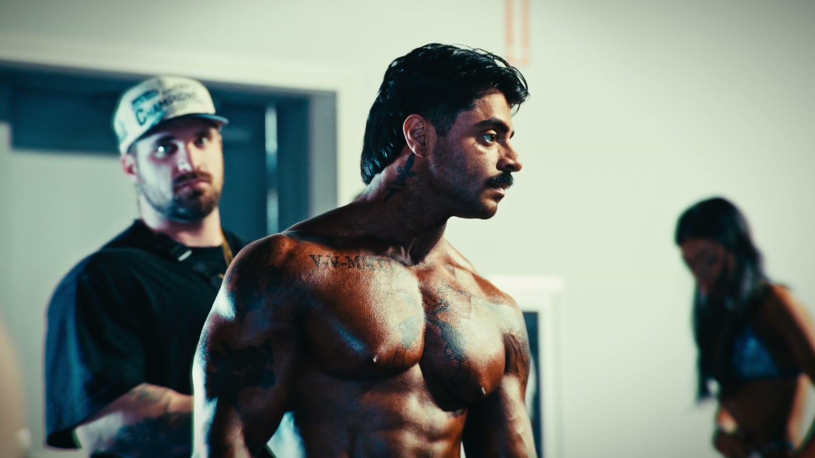 A tattooed bodybuilder backstage at a competition, looking off to the side, with a man in a white cap watching behind him