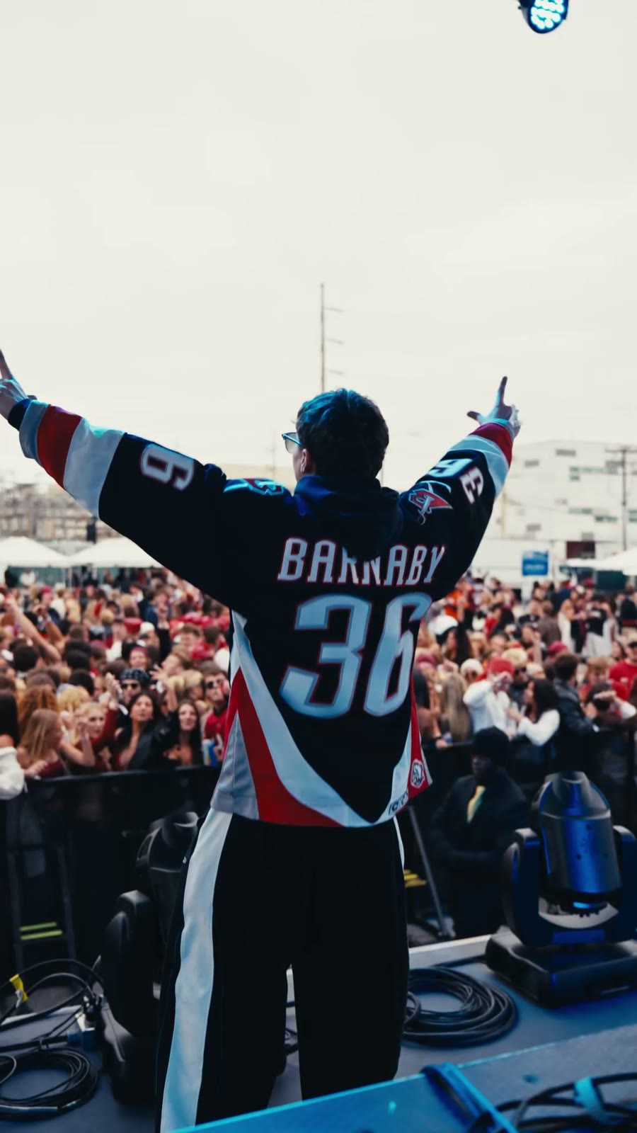 A performer in a number 36 jersey on stage with his arms raised, facing a packed outdoor crowd under a grey sky