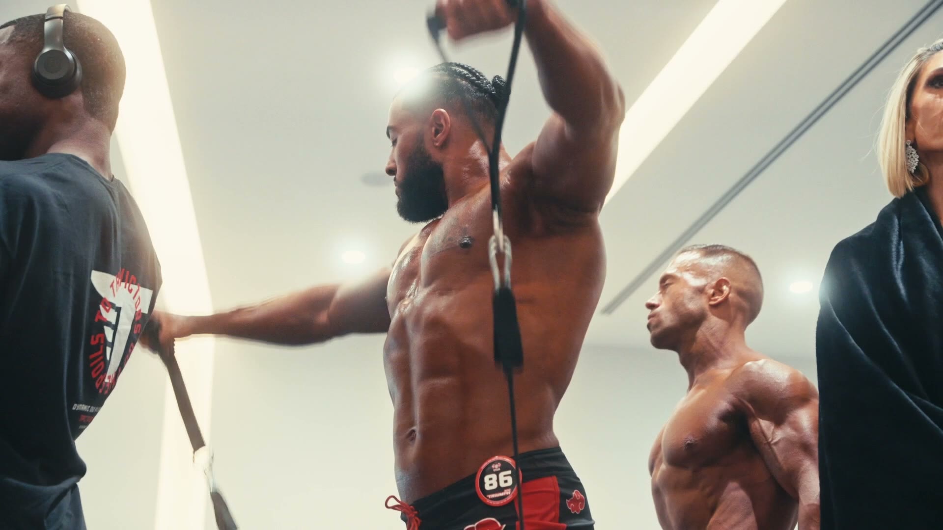 Low-angle shot of a bodybuilder wearing competitor number 86 warming up with a resistance band backstage, with another competitor flexing beside him