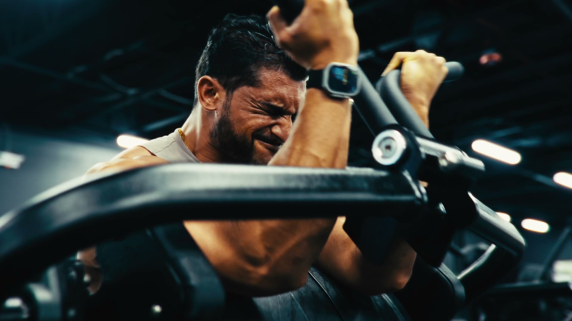 Close-up of a bearded man in a sleeveless shirt gritting his teeth through a curl on a gym machine, with warm lights blurred behind him