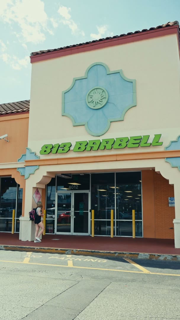 The front of the 813 Barbell gym on a sunny day, green letters on a cream wall above glass doors, a woman with a backpack walking toward the entrance