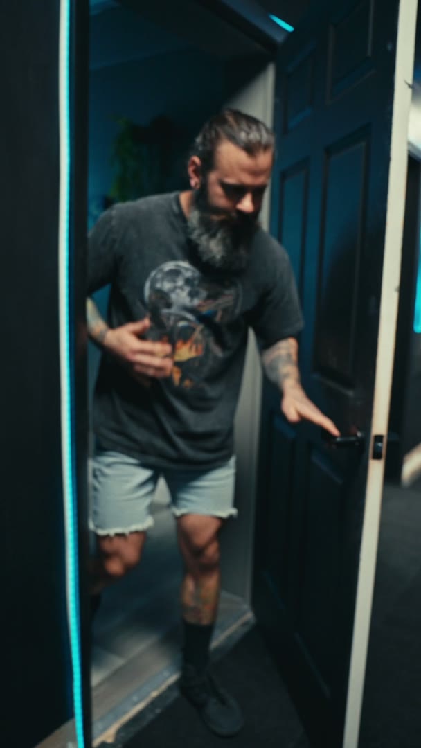 A bearded man in a gray graphic T-shirt and denim shorts steps out through a dark office door lit by a blue LED strip