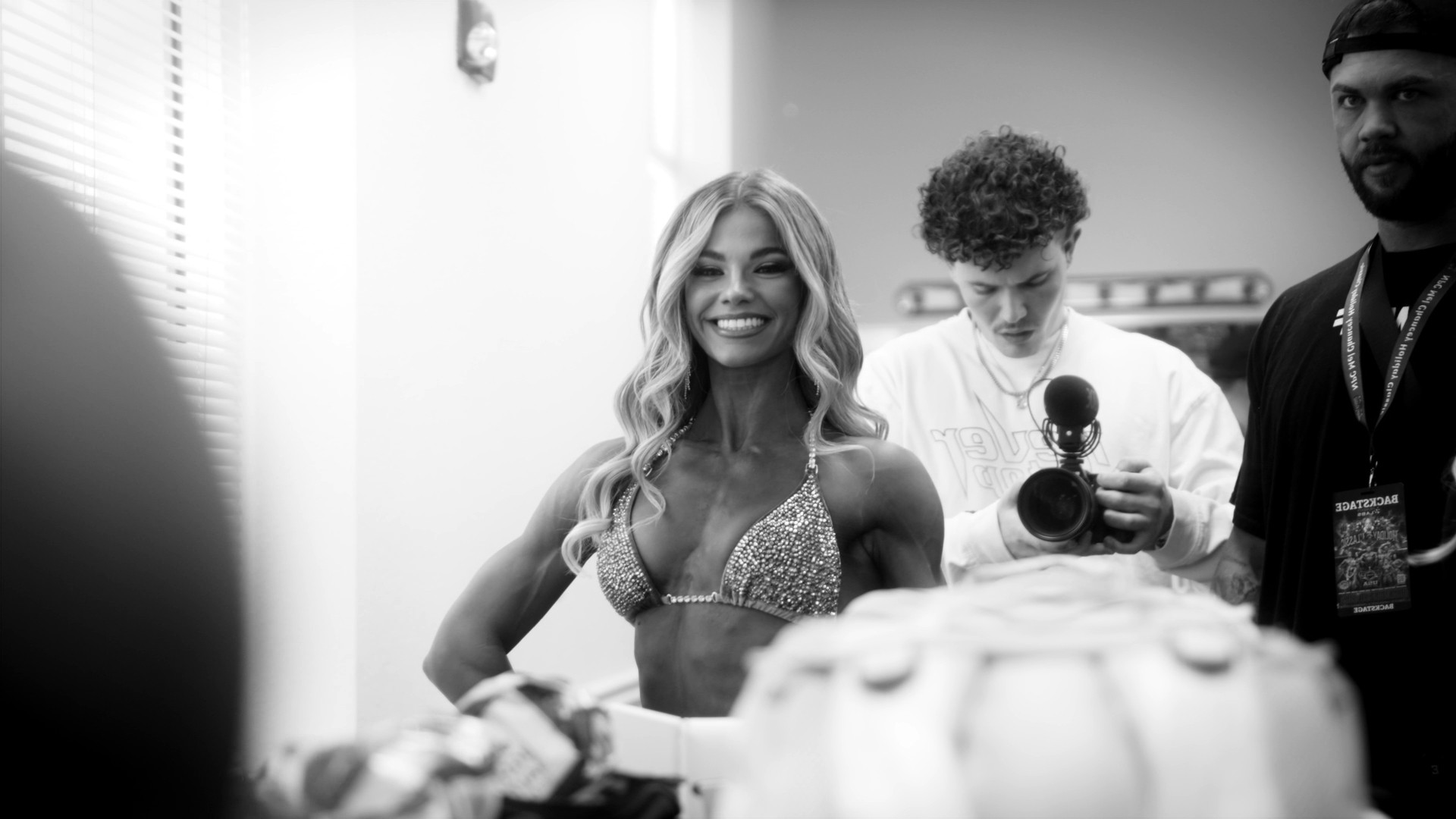 Backstage at a bodybuilding show, a competitor smiling while Grayson films
