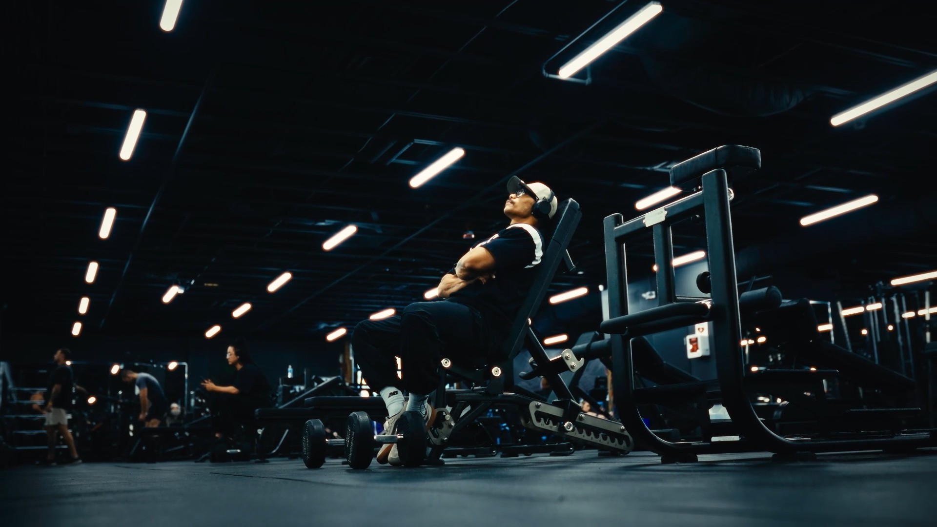 A man in headphones resting on an incline bench under the strip lights of a dark gym floor